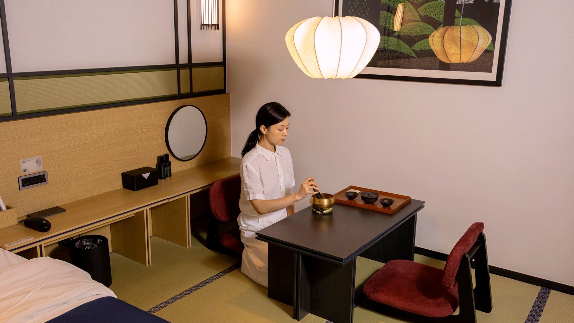 A hotel room with a bed, a desk with a computer, a chair, a small table with a tea set, and a wall-mounted mirror.