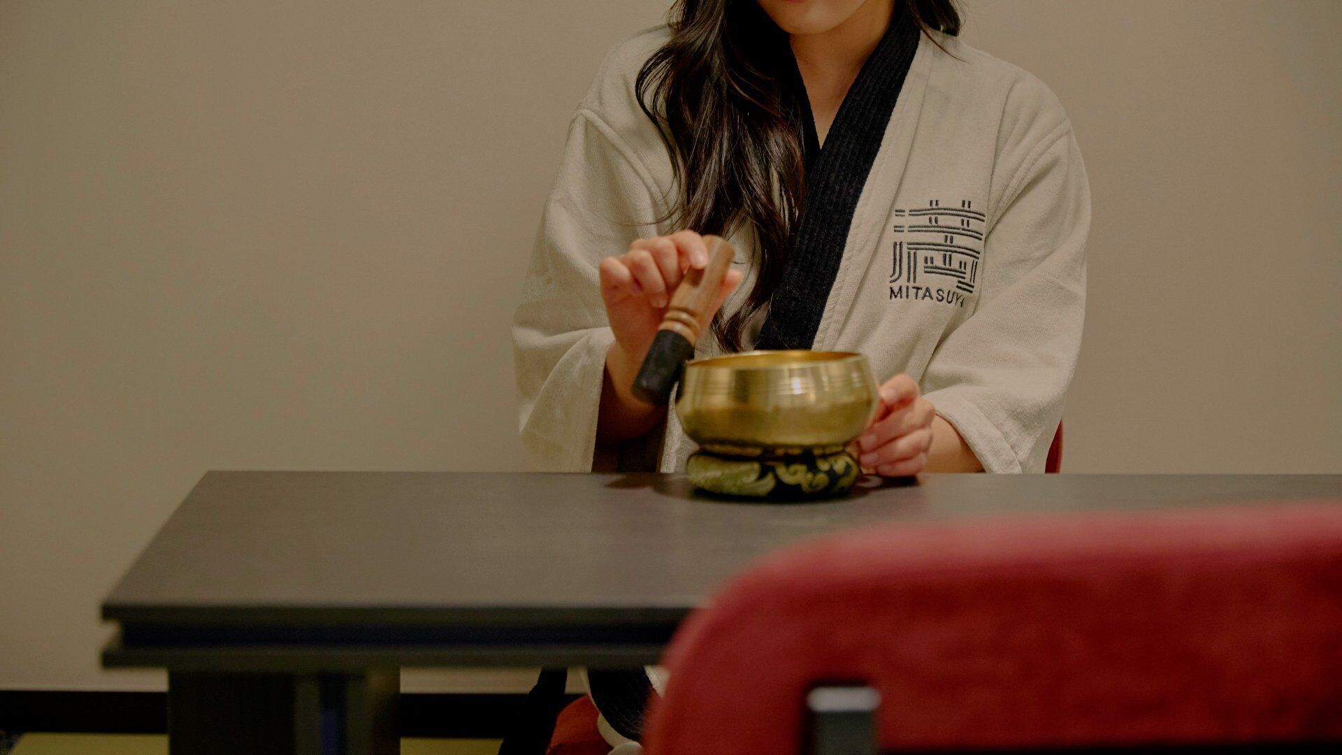 A person in a white robe sitting at a table with a bowl and a mallet, suggesting a tea ceremony setting.