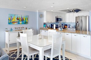 A modern kitchen with white cabinetry, a stainless steel refrigerator, and a microwave. There is a dining area with a white table and chairs.