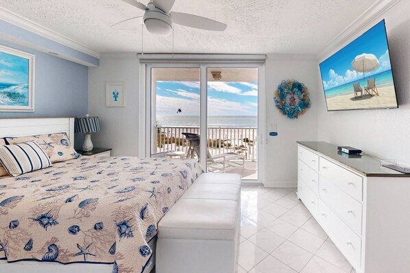 A bedroom with a bed, a bench, a dresser, and a balcony with a view of the beach.