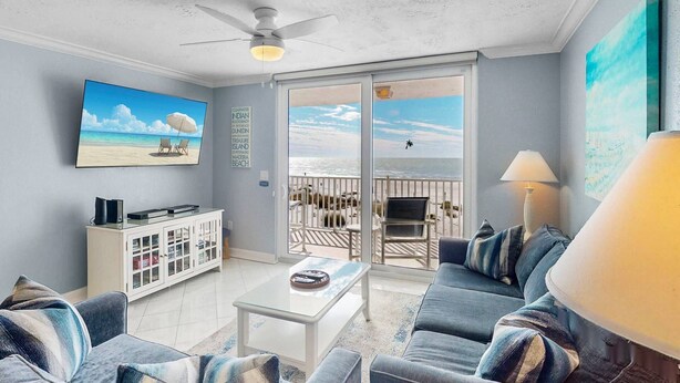 Een woonkamer met een plafondventilator, een televisie, een kast, een bank, een bijzettafeltje en een balkon met uitzicht op het strand.