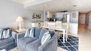 Een moderne woonkamer met een grijze zithoek, een eethoek met witte stoelen, een keuken met witte kasten en een blauw patroonkleed.