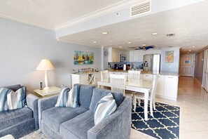 A modern living room with a grey sofa set, a dining area with white chairs, a kitchen with white cabinets, and a blue patterned rug.
