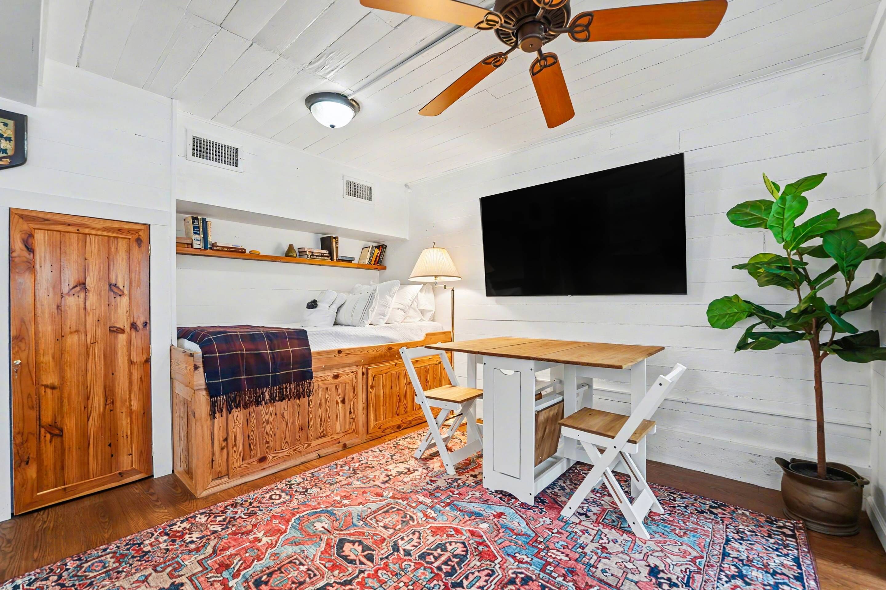 Una habitación acogedora con cama, escritorio, silla, ventilador de techo, televisor, estanterías y una planta en maceta.