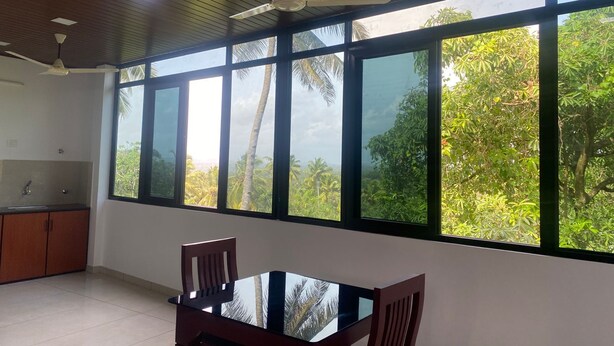 A room with a dining table and chairs, a kitchen area, and large windows with a view of greenery.