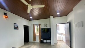 A spacious living area with a sofa, a flat-screen TV mounted on the wall, a ceiling fan, and a tiled floor.