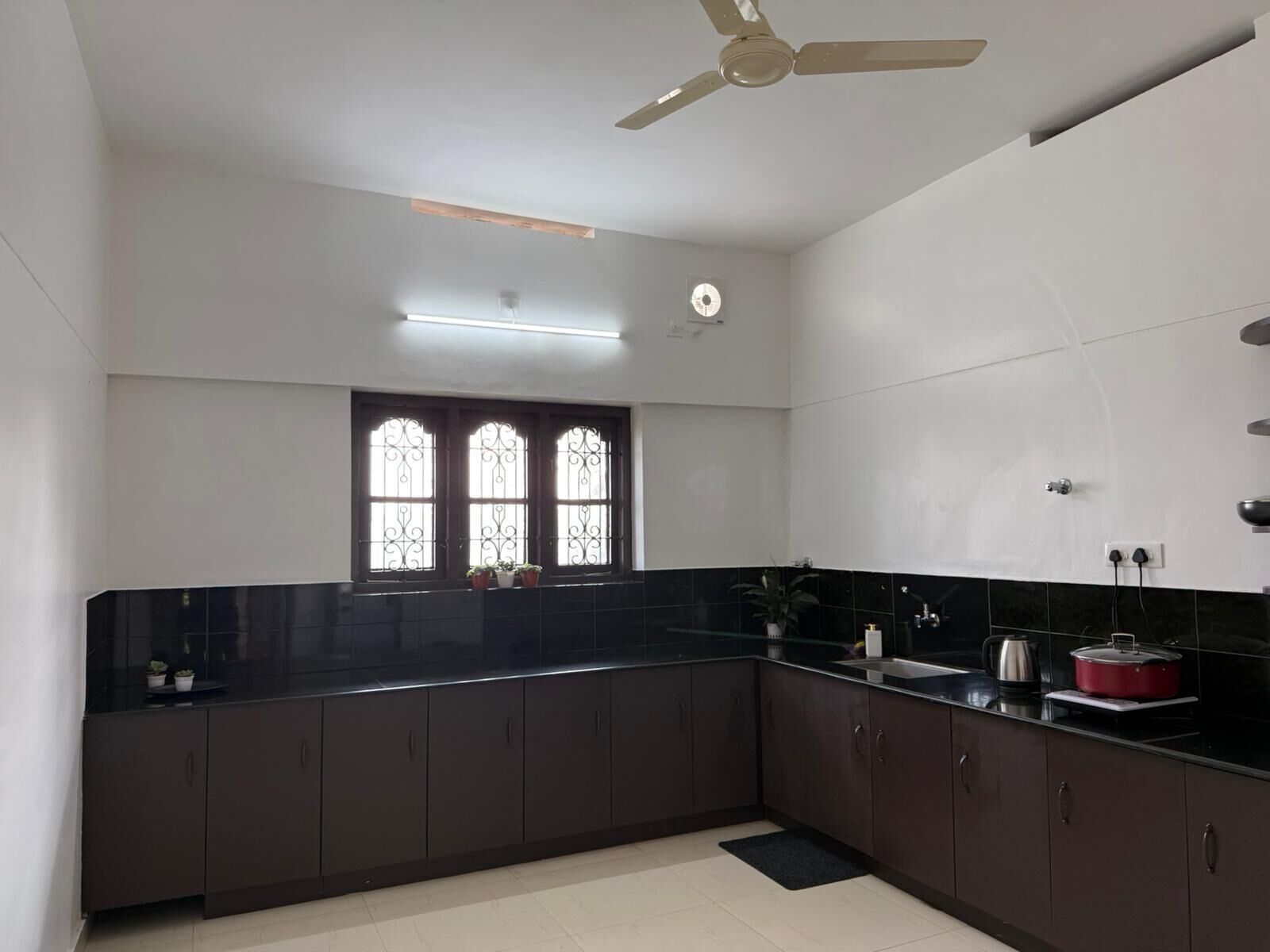 A modern kitchen with dark cabinets, a black countertop, a ceiling fan, and a window with decorative iron bars.