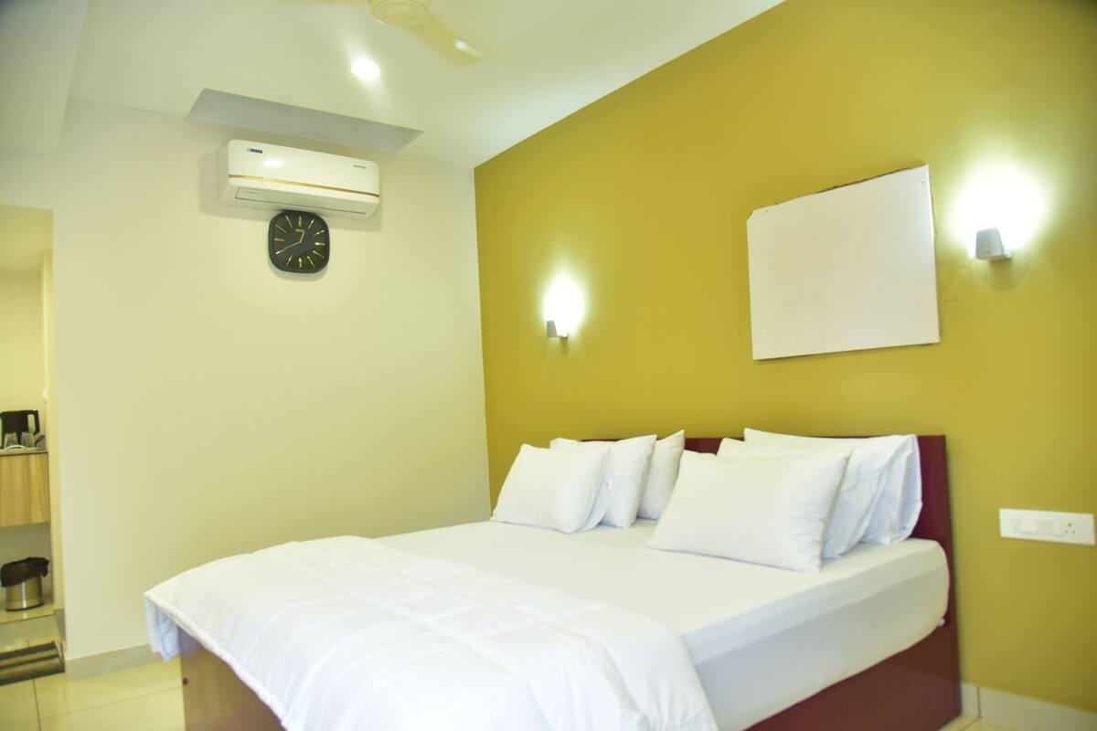 A hotel room with a bed, a wall clock, an air conditioning unit, and a light switch.