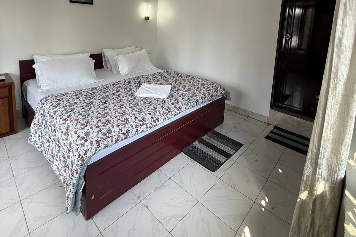 A bedroom with a bed, a nightstand, a wall-mounted light, a painting, and a door.