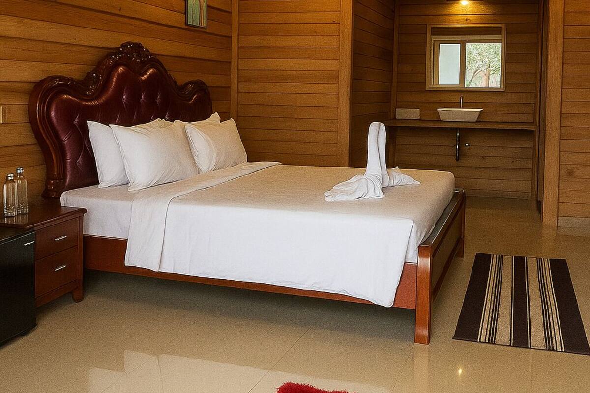 A wooden-paneled bedroom with a large bed, a nightstand, a red rug, and a window with a view of trees.