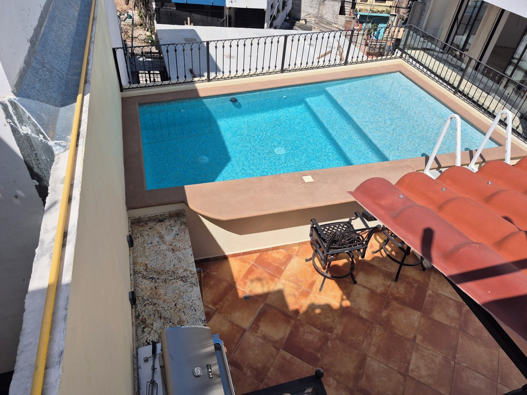Une piscine sur le toit, avec une eau d’un bleu limpide, entourée d’une rambarde en métal. On y trouve des chaises longues et une table sous un auvent rouge.