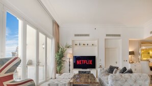 Un salón amplio con un gran ventanal, un sofá capitonado, una mesa de centro y un televisor que muestra 'NETFLIX'.