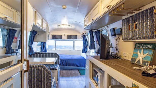 A compact RV interior with a kitchenette, a dining area, a sleeping berth, and a small entertainment unit.