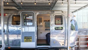 A vintage Airstream trailer with a covered porch, a bench, and a small interior space with a window and a door.