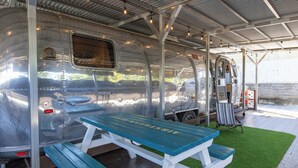 A vintage trailer with a picnic table and benches, a covered area with a metal roof, and a grassy area outside.
