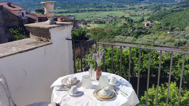 A round table set for two with white tablecloth and plates, a vase with flowers, and a view of green hills and mountains in the background. - Historic home, wonderful view of the hills, 20" from the Royal Palace of Caserta. (Caiazzo)