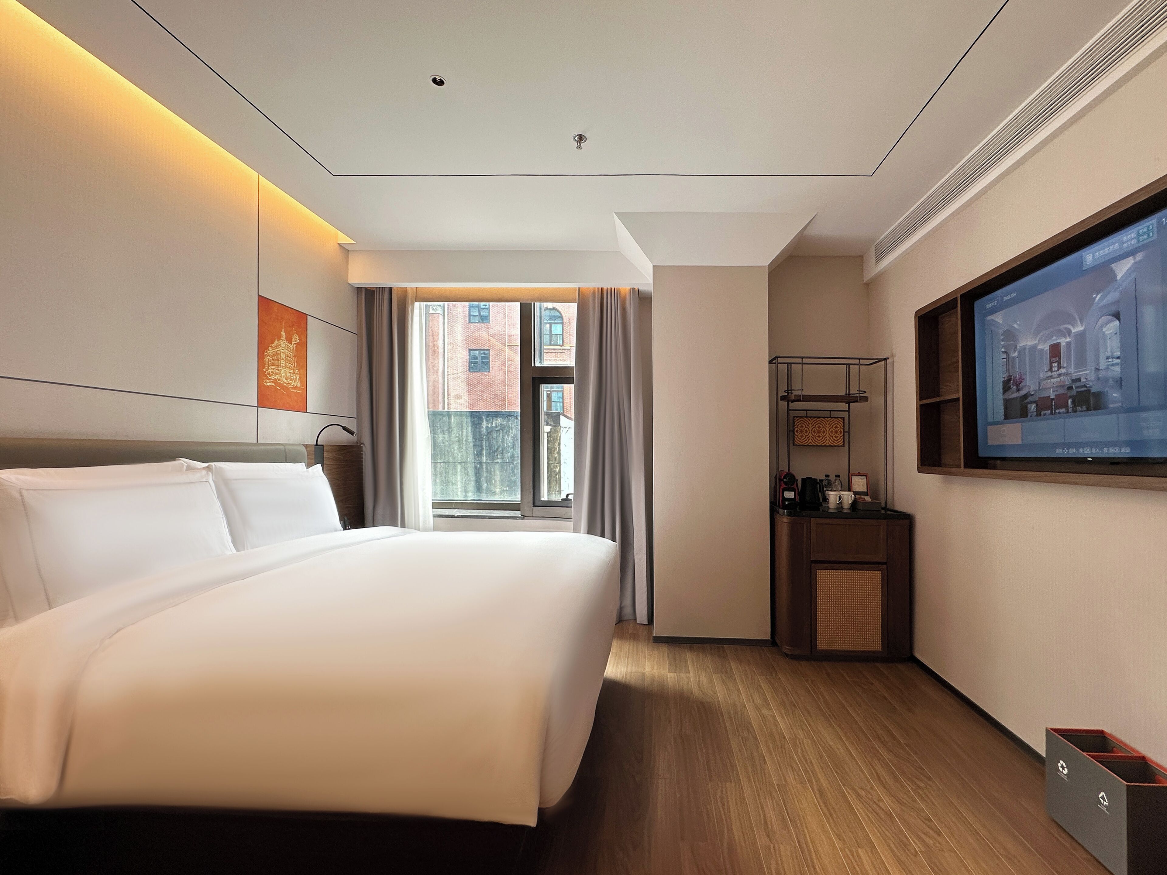 A hotel room with a large bed, a television mounted on the wall, and a window with a view of buildings outside.