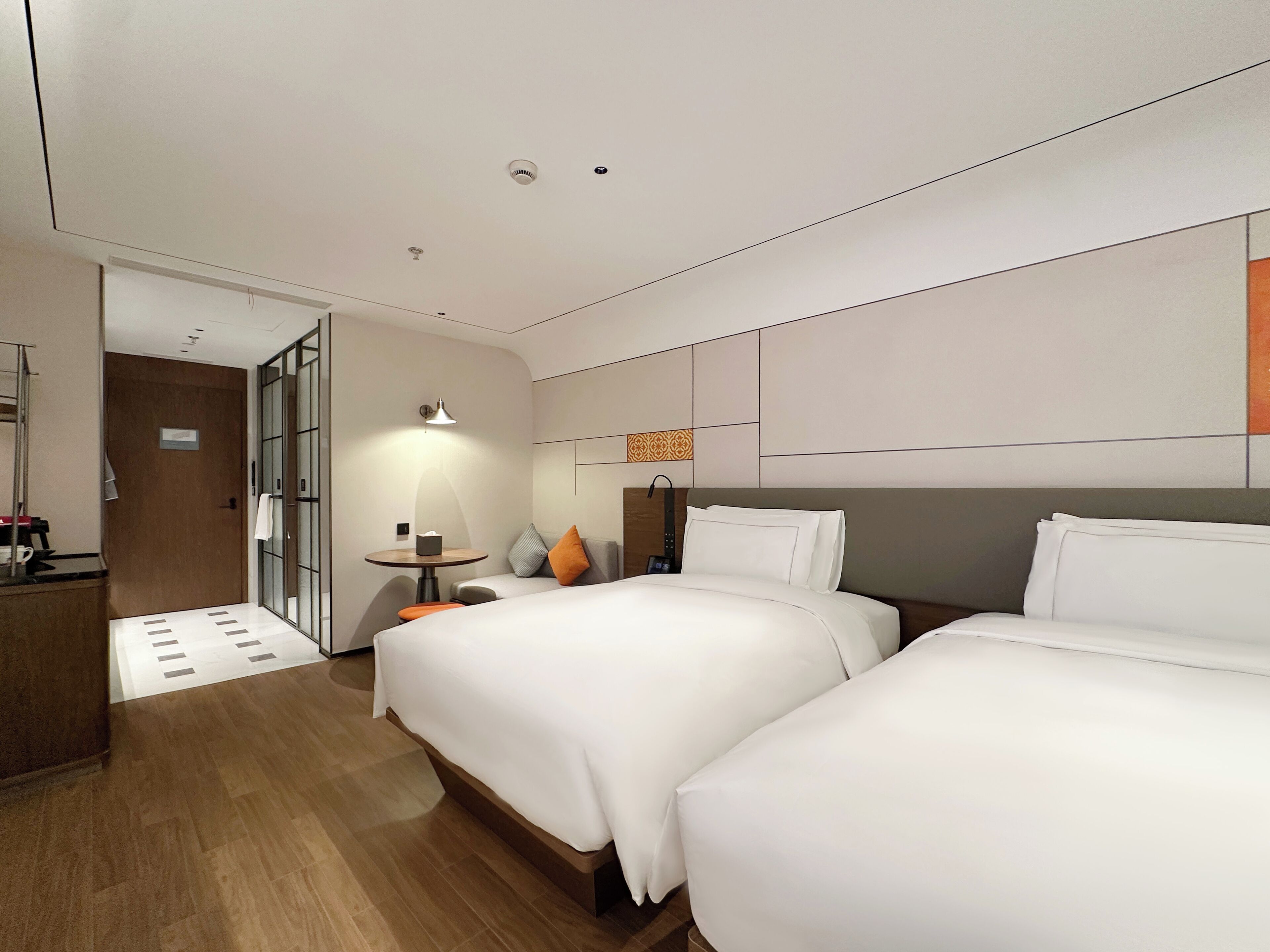 A modern hotel room with two beds, a desk, a chair, a small table with a lamp, and a bathroom visible through an open door.