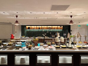Disposición de bufé con diversos alimentos y utensilios, una barra con caja registradora y una pared decorativa con diseño verde.