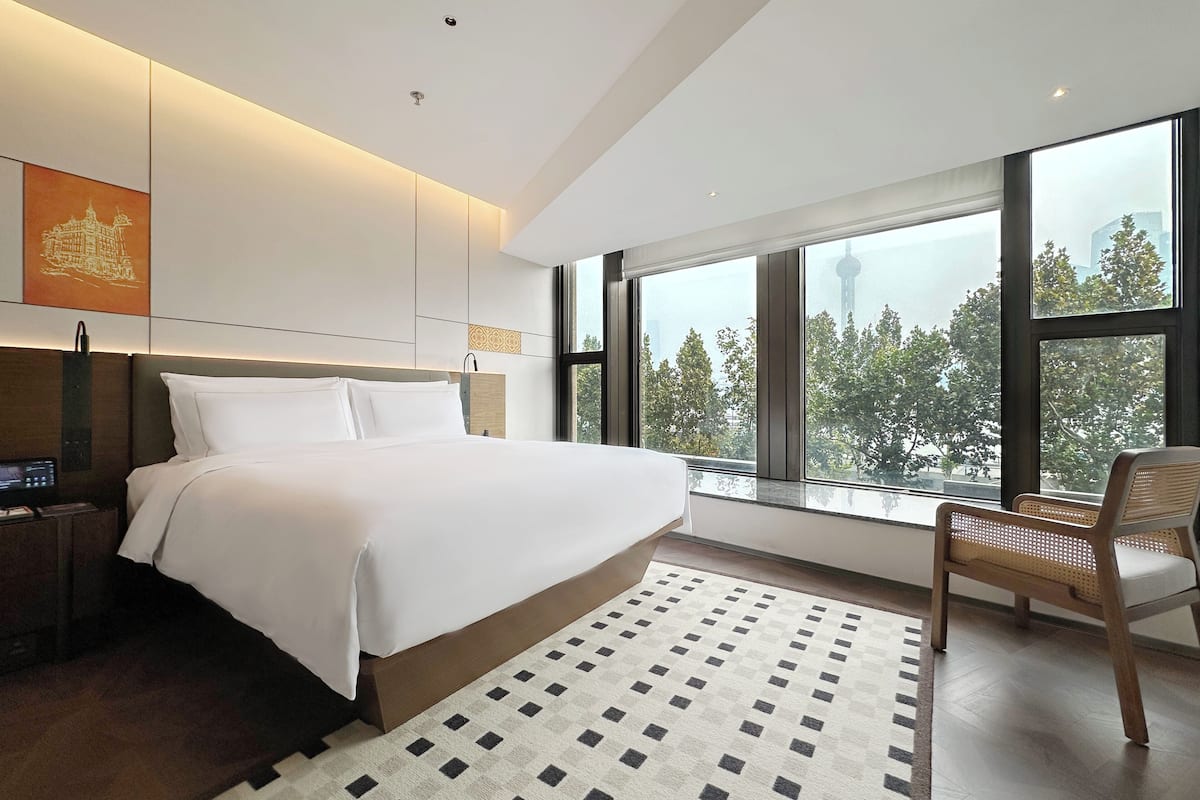 Une chambre d’hôtel moderne avec un grand lit, une table de chevet en bois, une chaise et une vue sur des arbres et une tour, le tout visible à travers de grandes fenêtres.