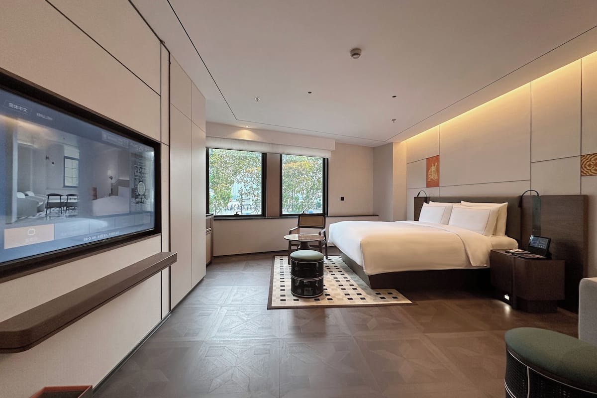 Une chambre d’hôtel moderne, dotée d’un grand lit, d’un bureau, d’une chaise, d’une télévision et d’une fenêtre donnant sur un paysage arboré.