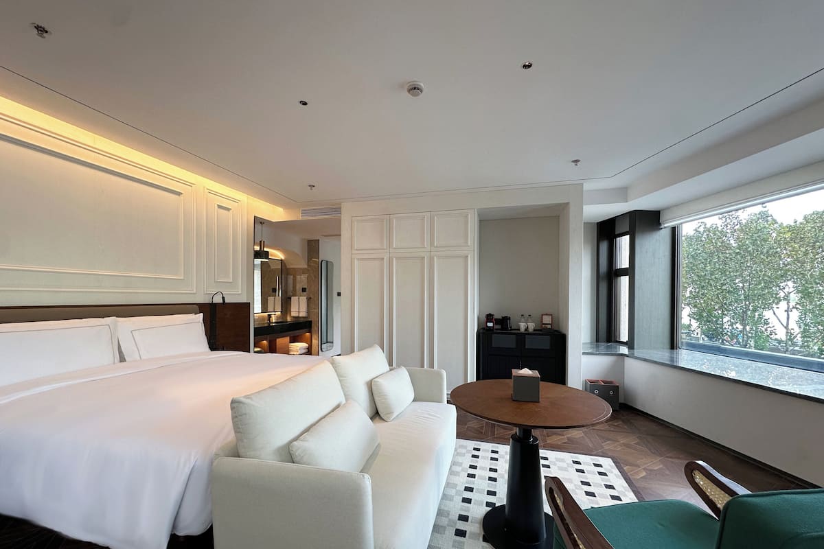 Une chambre d’hôtel avec un grand lit, un canapé, une petite table et une vue sur les arbres par la fenêtre.