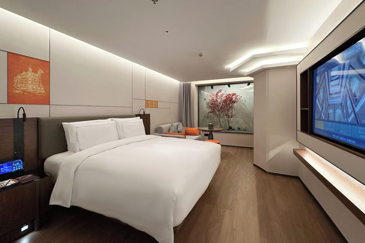 Une chambre d’hôtel moderne dotée d’un grand lit, d’un bureau avec un ordinateur portable, d’une télévision et d’une fenêtre donnant sur un immeuble et un arbre.