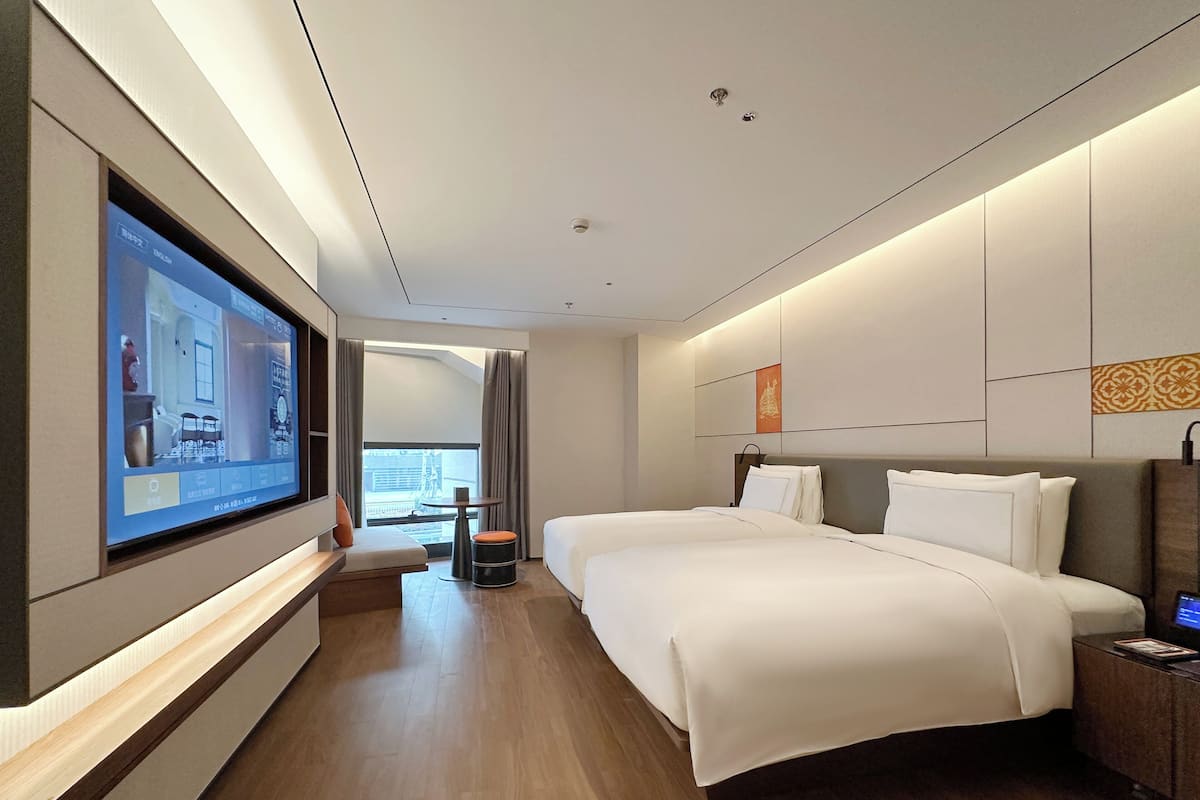 Une chambre d’hôtel moderne dotée d’un grand téléviseur à écran plat fixé au mur, d’un lit avec du linge de maison blanc, d’une petite table ronde avec une chaise, et offrant une vue sur l’extérieur grâce à une grande fenêtre.