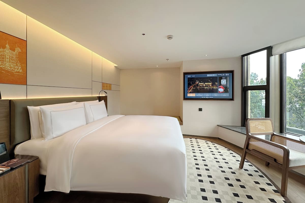Une chambre d’hôtel moderne dotée d’un grand lit, d’un téléviseur à écran plat fixé au mur, d’un coin salon avec un fauteuil et d’une vue sur l’extérieur grâce à une grande fenêtre.