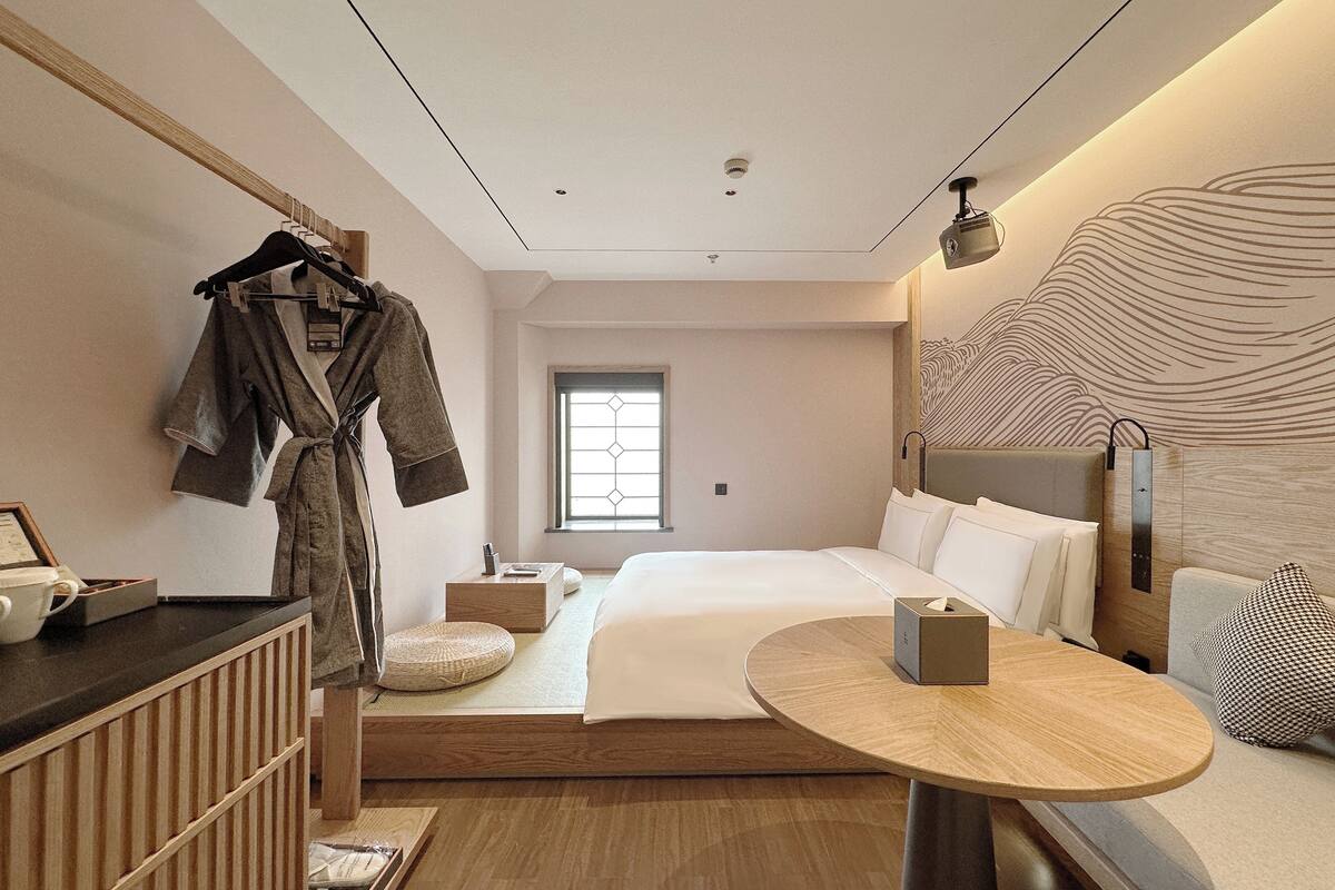 Une chambre d’hôtel moderne avec un grand lit, une table de chevet, un peignoir suspendu à un porte-manteau et une vue sur l’extérieur par une fenêtre.