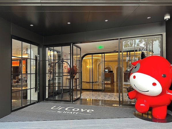L’entrée d’un hôtel, avec une grande mascotte rouge, des portes en verre et une enseigne indiquant « Ur Cove by Hyatt ».