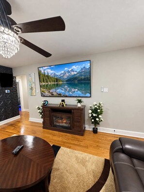 A living room with a fireplace, a television mounted on the wall, a ceiling fan, and a dining area with a table and chairs.