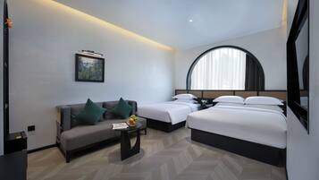 A modern hotel room with a sofa, two single beds, a small table with fruit, a framed picture, and a large window with curtains.