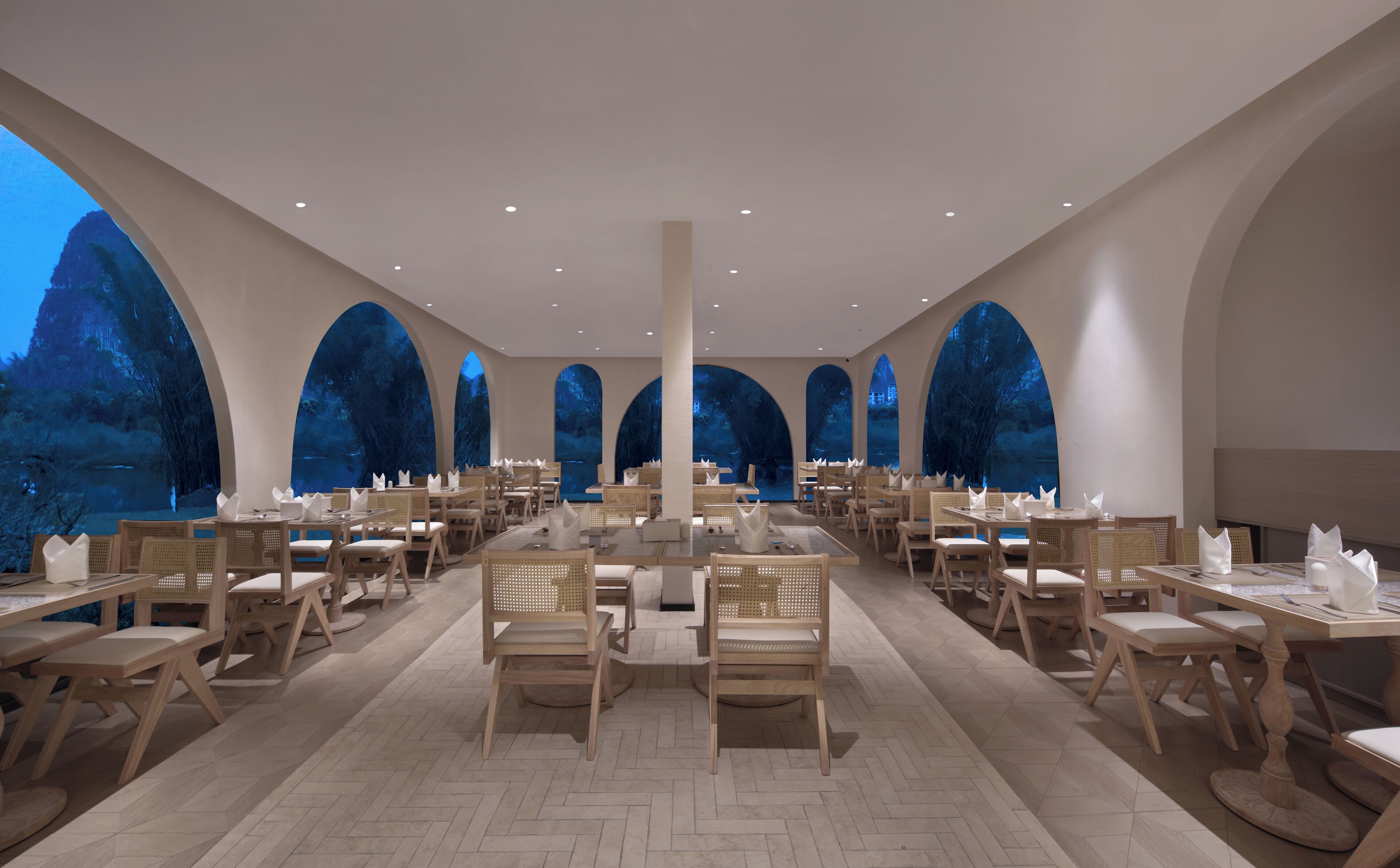 A spacious dining area with multiple tables and chairs, large arched windows, and a herringbone floor pattern.