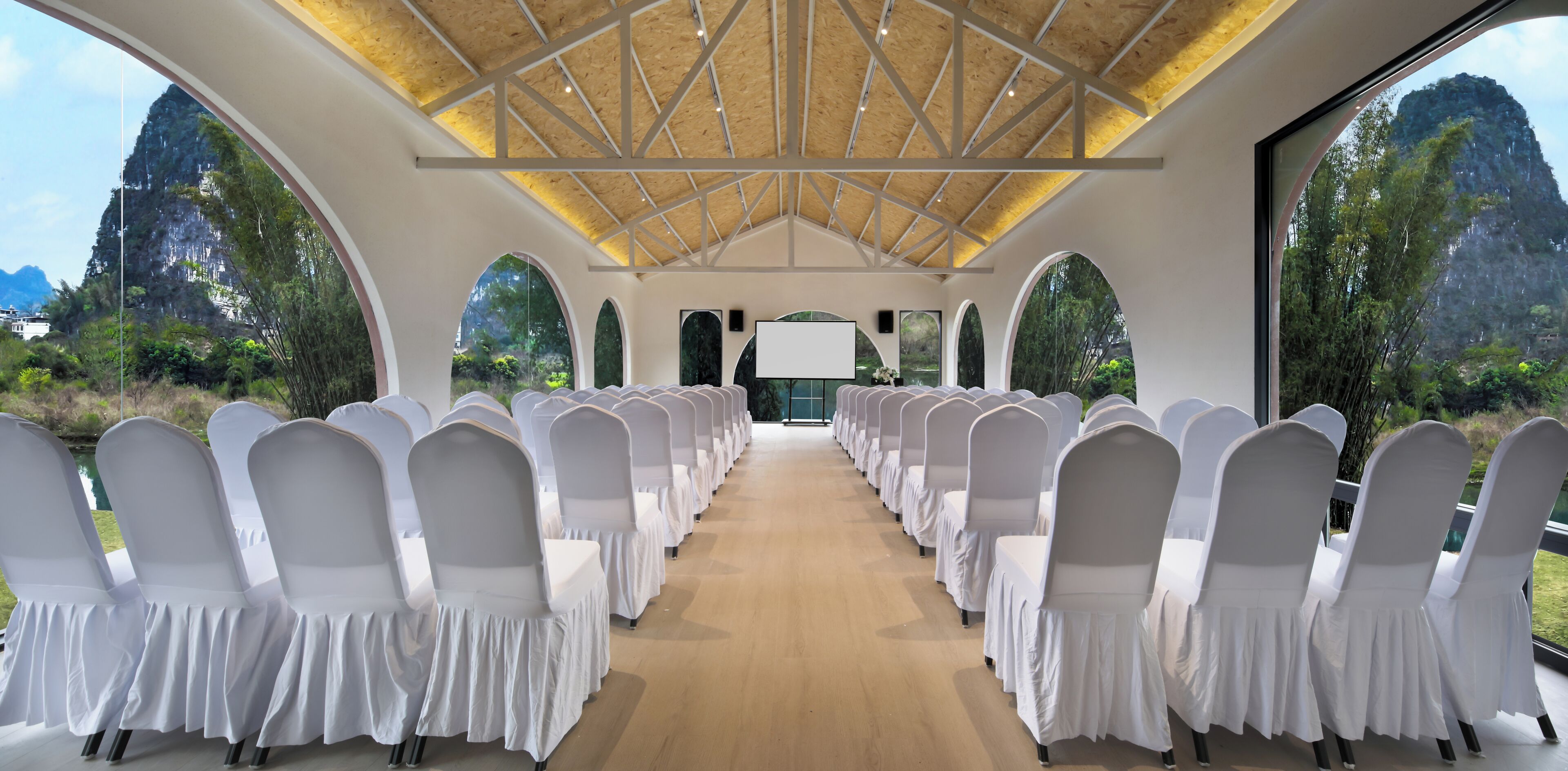 Una sala conferenze con file di sedie ricoperte di bianco rivolte verso un grande schermo, con vista su un paesaggio naturale e un soffitto a struttura in legno.