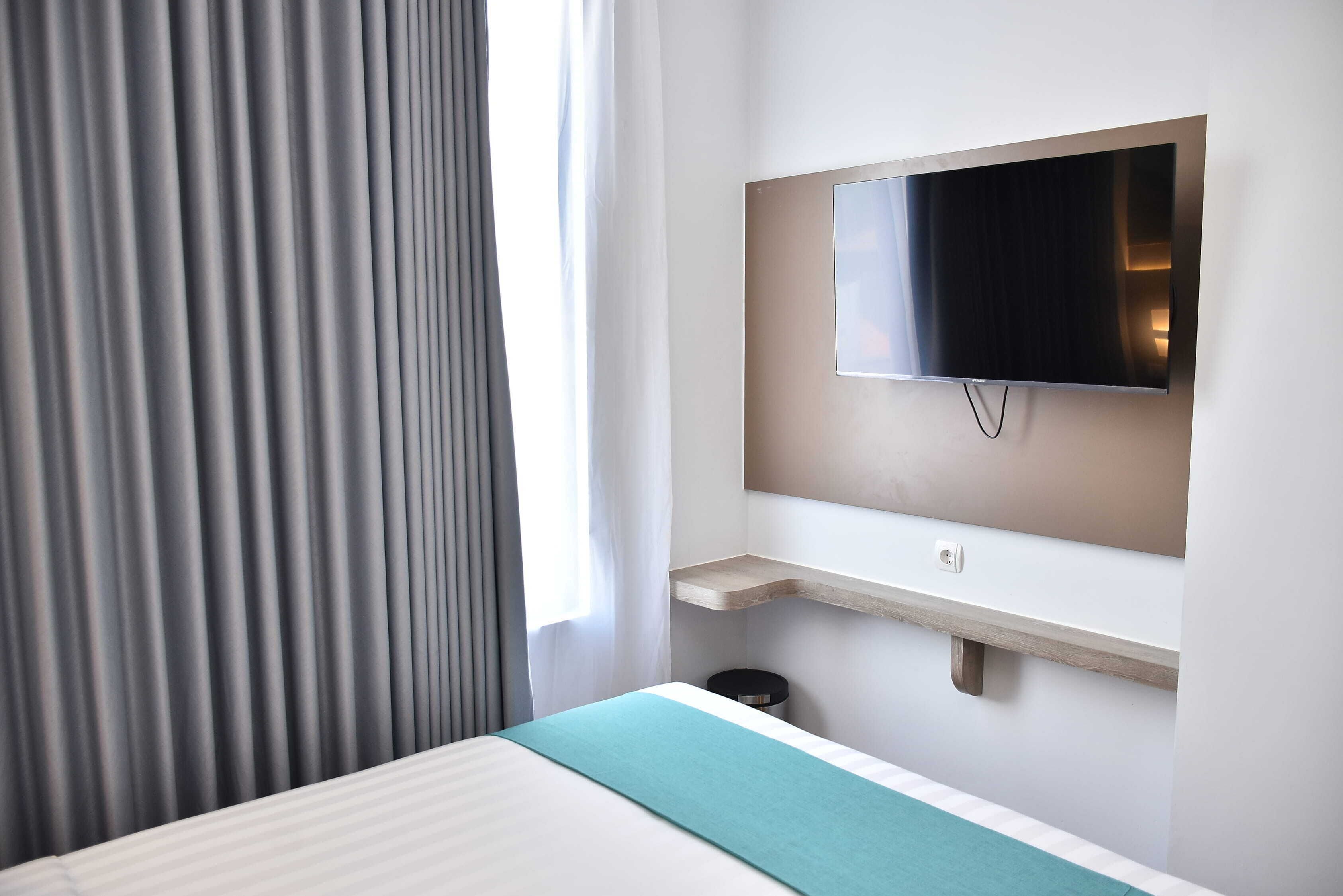 Ein Hotelzimmer mit einem Bett, einem an der Wand montierten Fernseher, einer kleinen Holztrennwand und einem Fenster mit Vorhängen.