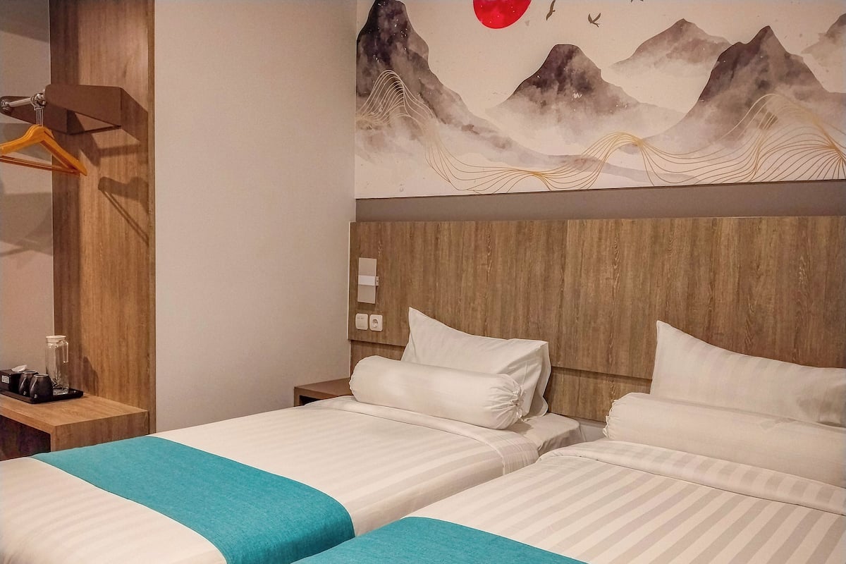 A hotel room with two beds, a wooden headboard, a painting of mountains, and a small table with a camera and a glass jar.