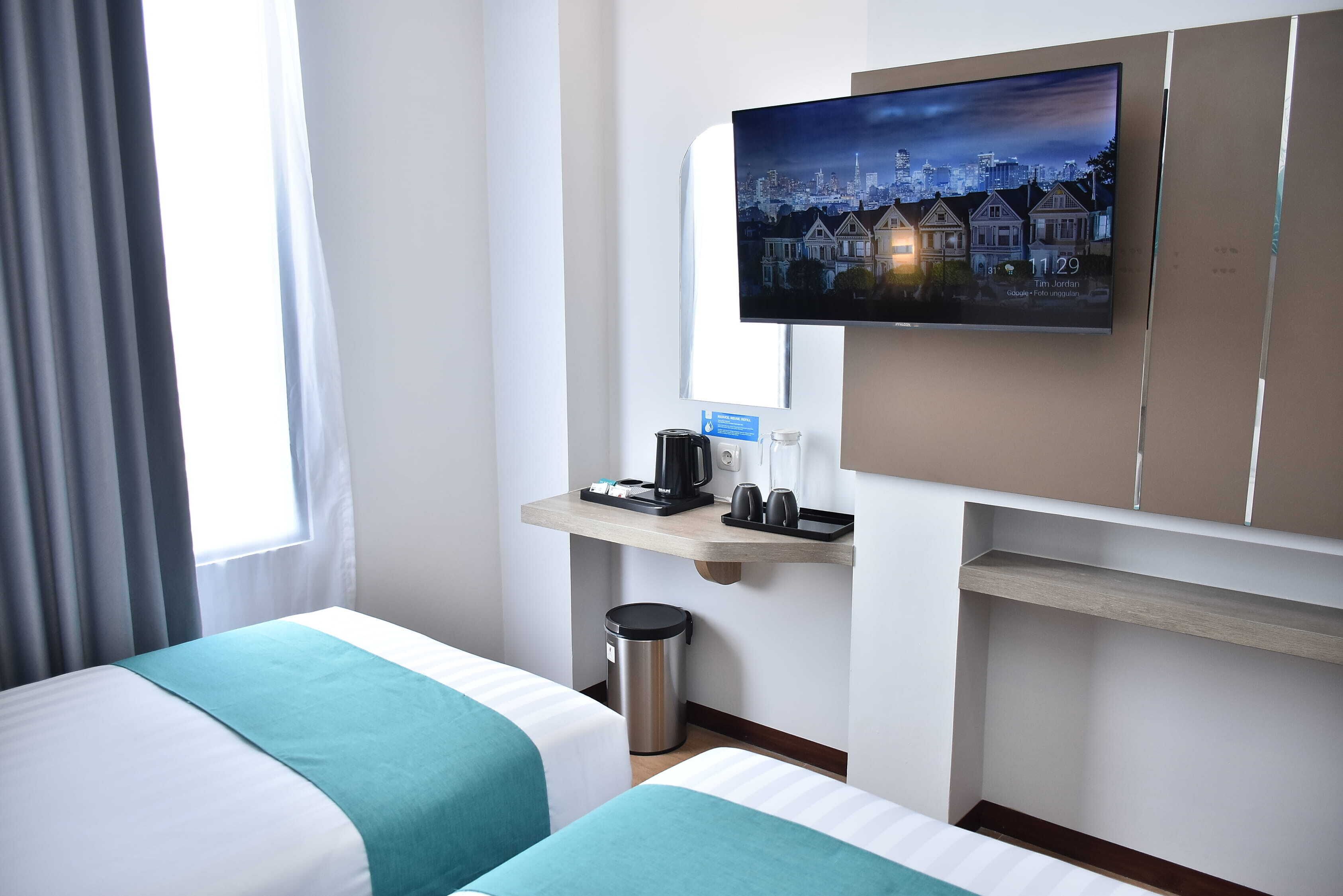 A hotel room with two beds, a TV mounted on the wall, a desk with a coffee maker, and a trash can.