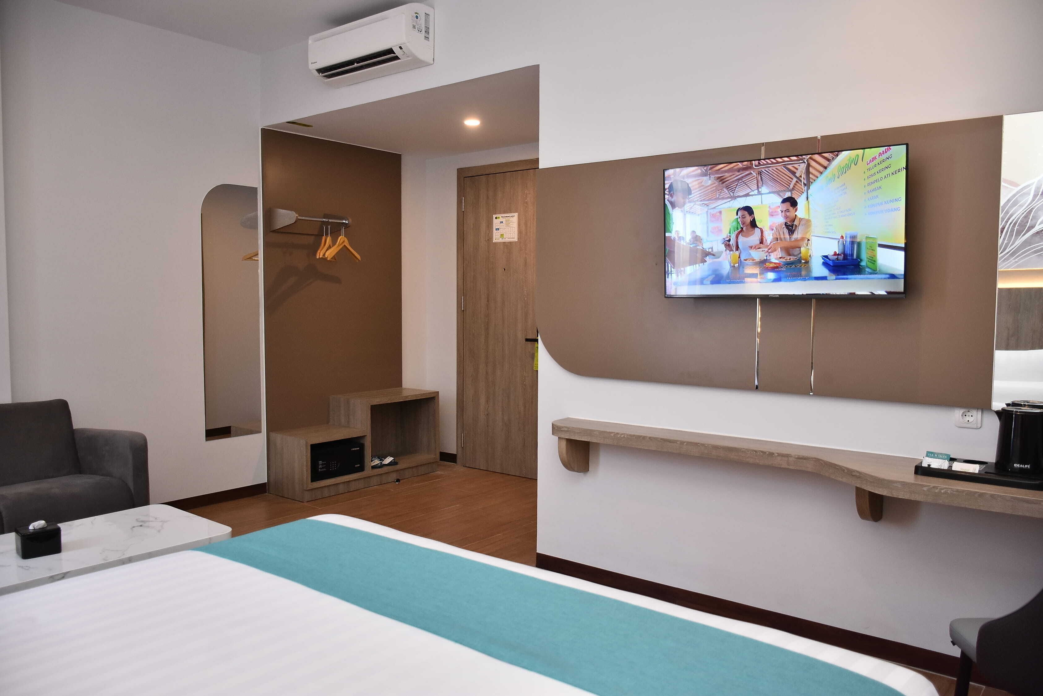 A hotel room with a bed, a TV mounted on the wall, a desk with a chair, and a wooden door.