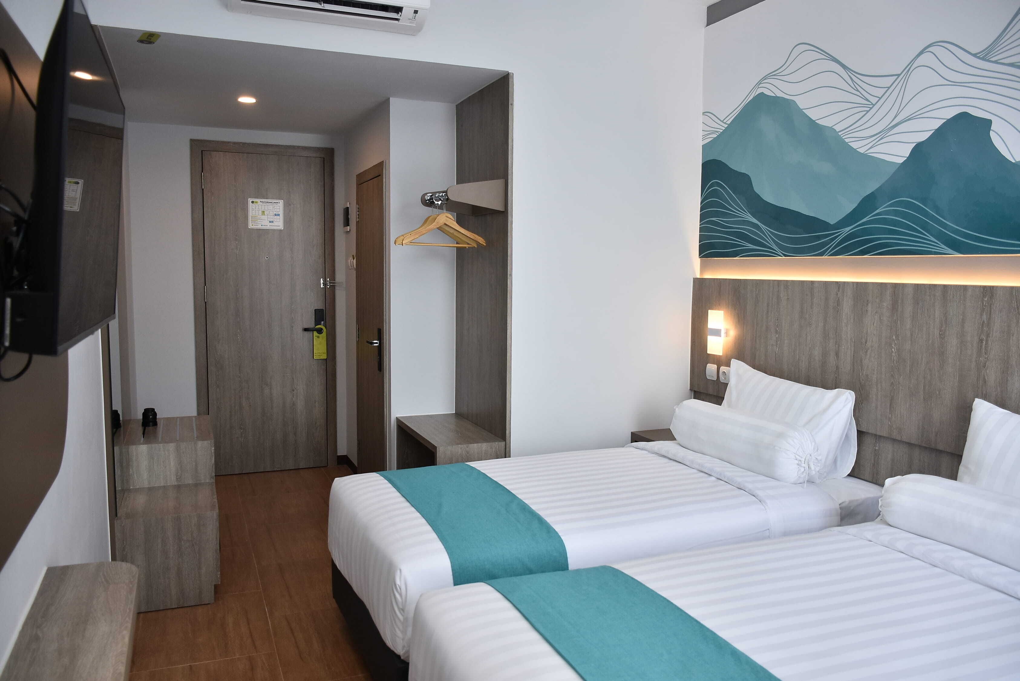 A hotel room with two beds, a wooden door, a coat hanger, a wall-mounted TV, and a painting of mountains.