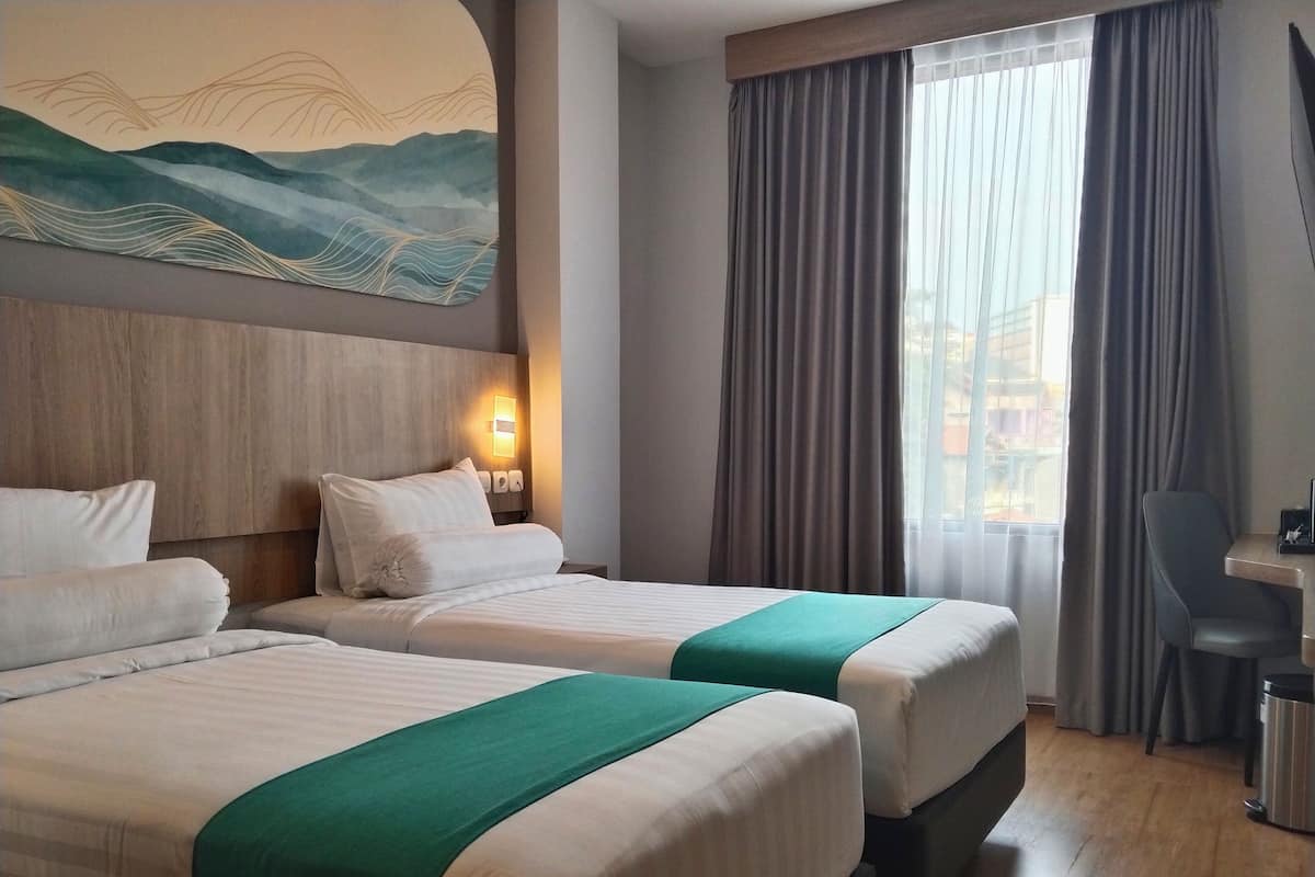 A hotel room with two beds, a desk, a chair, a painting on the wall, and a window with curtains.