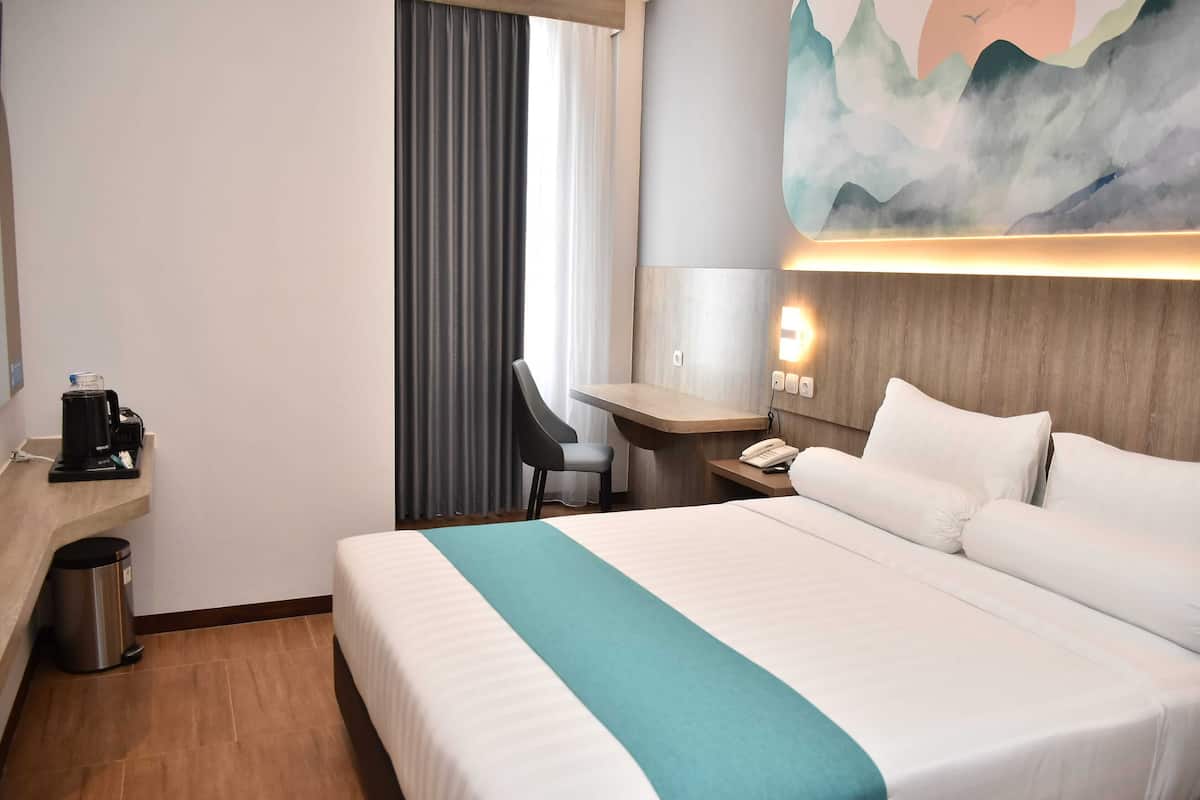 A hotel room with a bed, a desk, a chair, a coffee maker, a trash bin, and a painting on the wall.