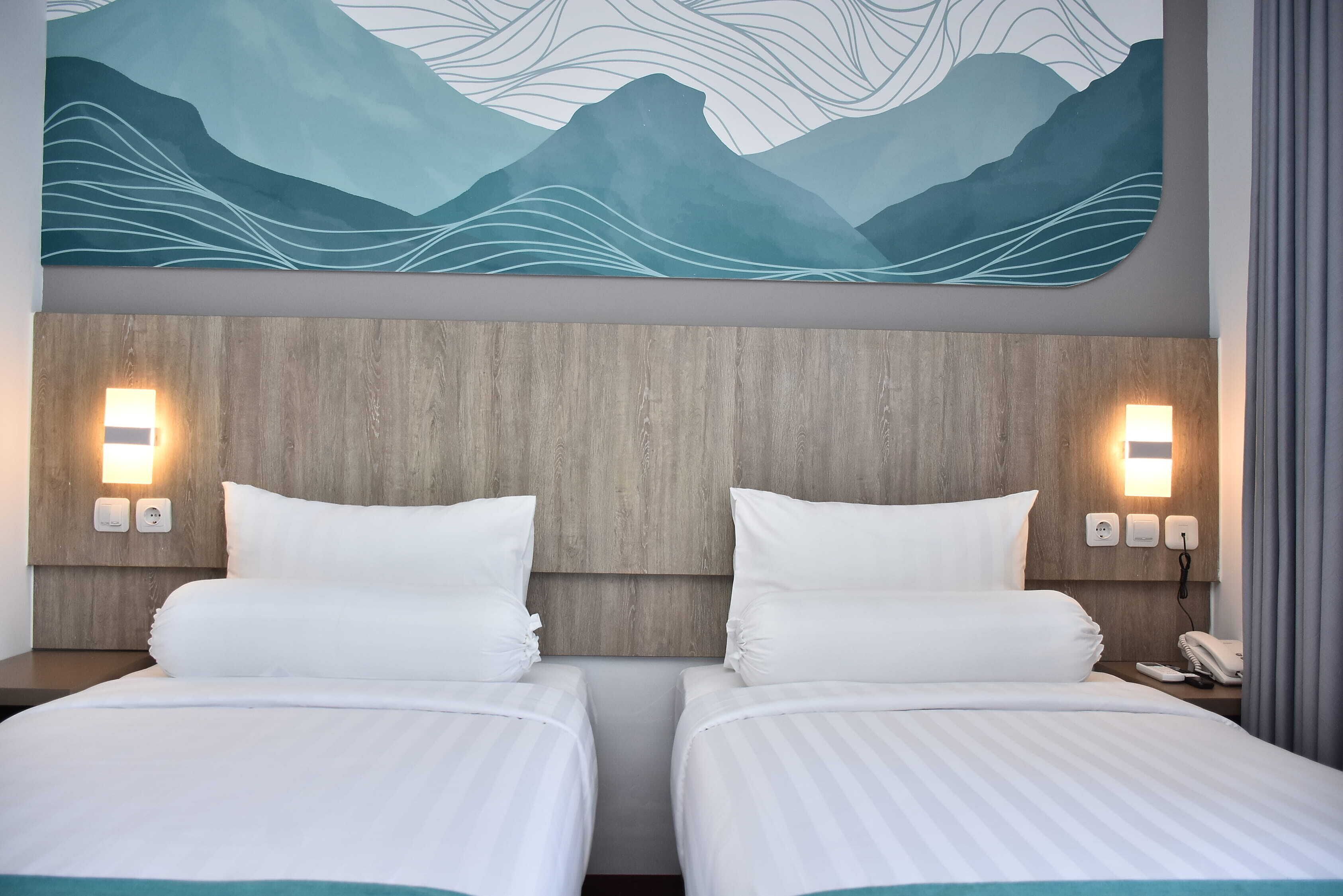 Habitación de hotel con dos camas, cabecero de madera, luces fijadas a la pared y un cuadro abstracto grande sobre las camas.