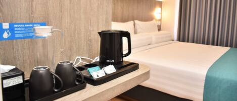 Habitación de hotel con una cama, una mesita de noche con hervidor, tazas y una bandeja con una bolsita de té y una tarjeta.