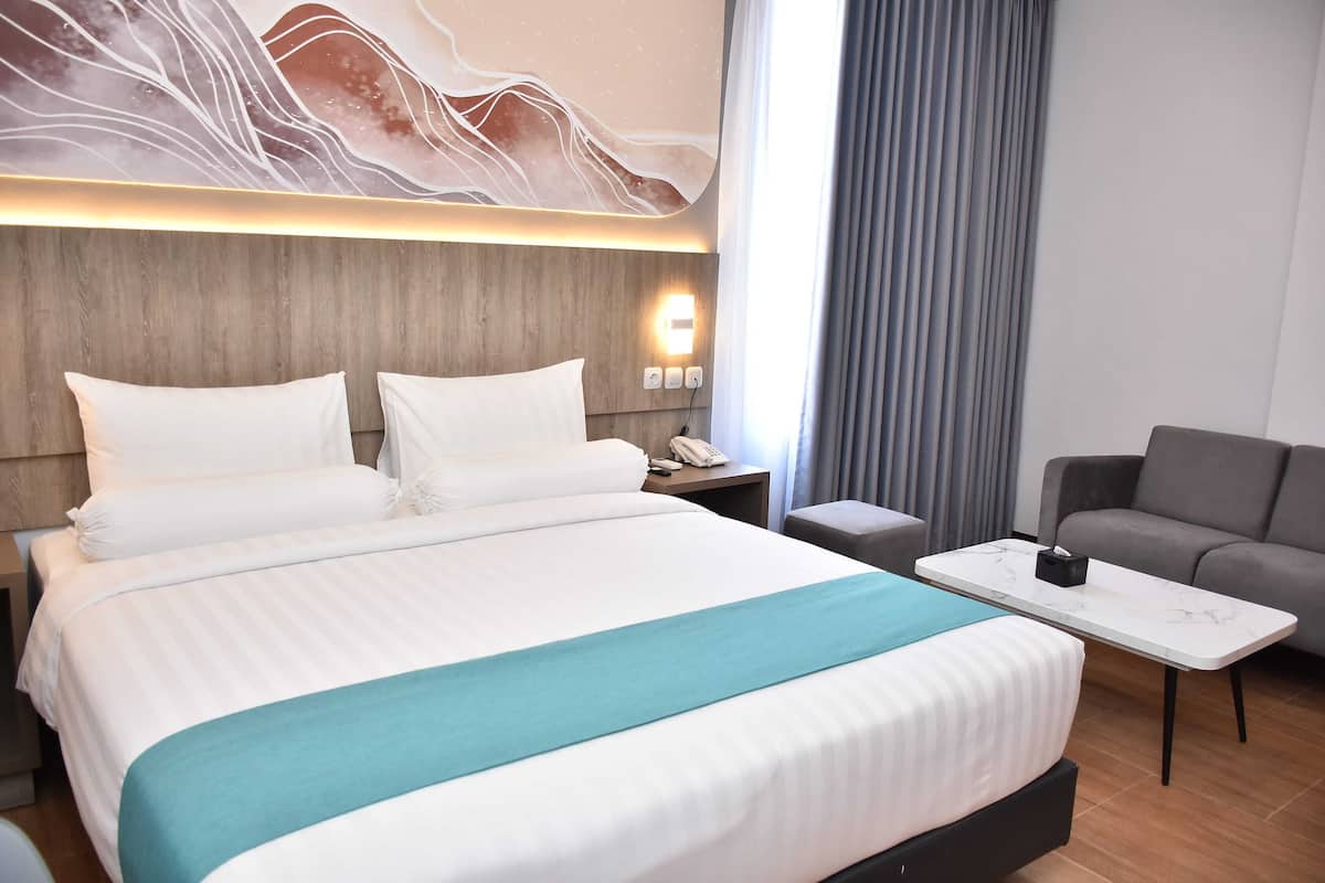 A hotel room with a large bed, a bedside table with a phone, a grey sofa, and a wall with a light fixture.