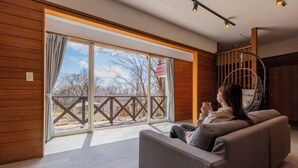 Un intérieur moderne avec un mur en bois, un canapé gris et une grande porte-fenêtre coulissante donnant sur un espace extérieur doté d’une terrasse en bois et offrant une vue sur les arbres.