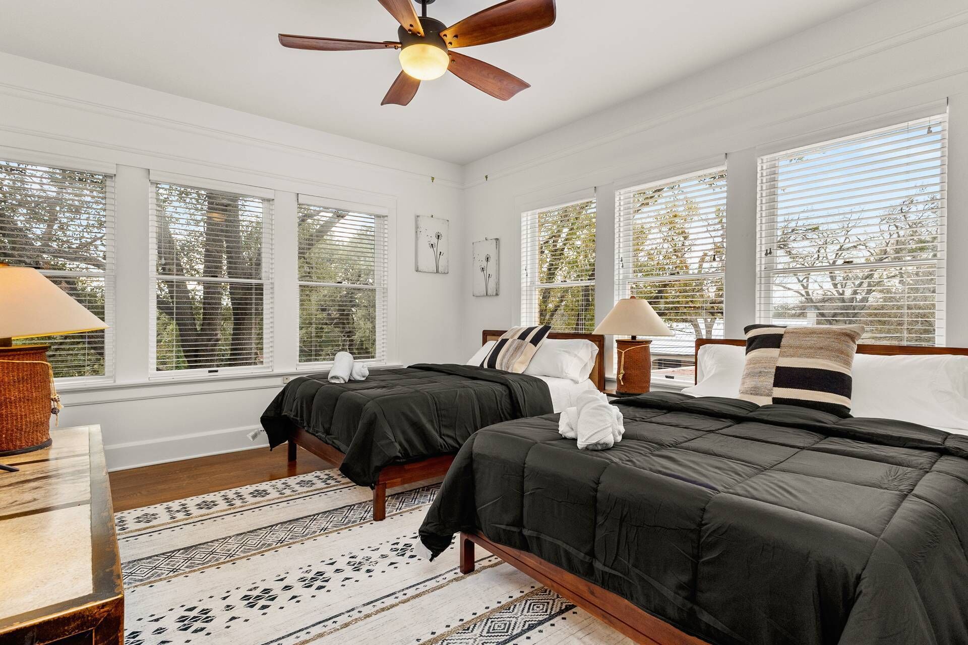 Une chambre avec deux lits, un ventilateur de plafond et de grandes fenêtres équipées de stores.