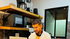 Una persona trabajando en una computadora portátil sobre un escritorio de madera en una cocina con microondas, cafetera y una ventana con persianas.