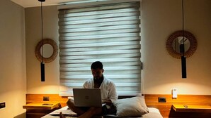 A person sitting on a bed in a hotel room with a laptop, a ceiling fan, and two wall-mounted lamps. - Jacaranda (Puerto Viejo de Talamanca)