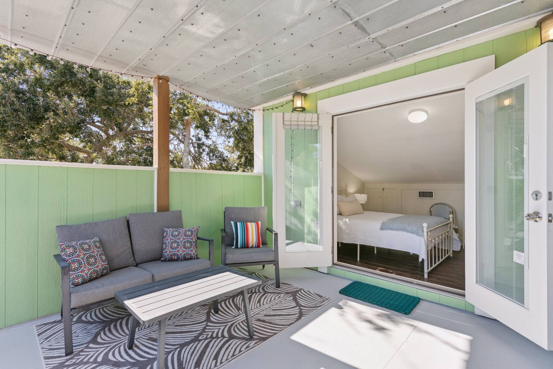 Un área de estar al aire libre acogedora, con un sofá y sillones, alfombra estampada y vista a un dormitorio con cama y ventana.